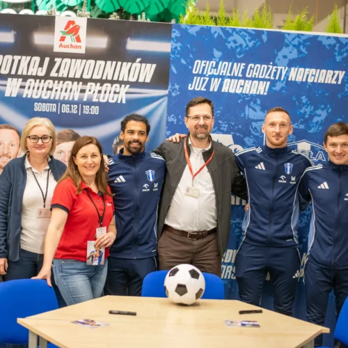Na zdjęciu siedem osób, w tym piłkarze w klubowych strojach oraz postać maskotki, pozują przed planszami reklamowymi w Auchan Płock podczas wydarzenia promującego zespół.