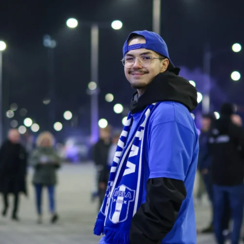 Mężczyzna w niebieskiej koszulce i czapce, z szalem w barwach klubu sportowego, uśmiecha się na tle nocnego miasta z jarzącymi się światłami.