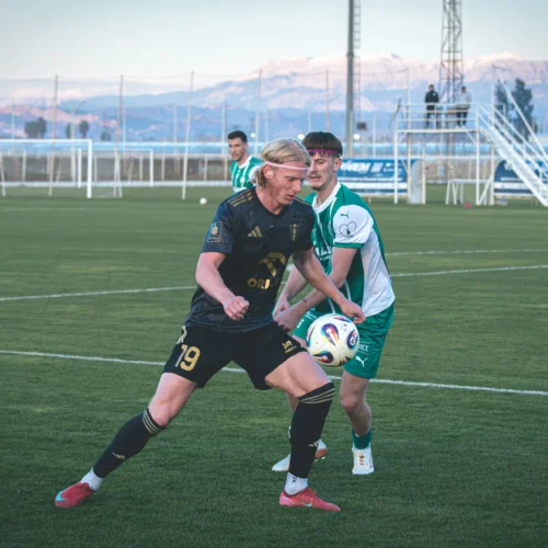 Na zdjęciu widać dwóch piłkarzy rywalizujących o piłkę na boisku, z widokiem na górskie tło.