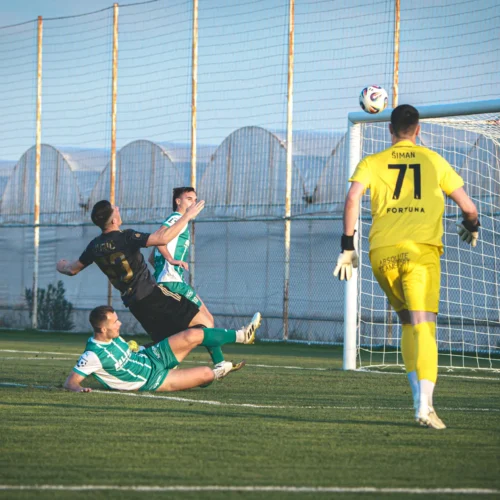 Na zdjęciu widać piłkarzy podczas meczu, gdzie jeden z zawodników próbuje oddać strzał na bramkę, a bramkarz jest gotowy do obrony.
