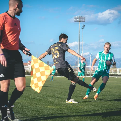 Na zdjęciu widać piłkarzy podczas meczu na boisku, z sędzią i asystentem na pierwszym planie.