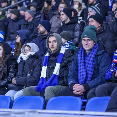 Na zdjęciu widać grupę kibiców siedzących na trybunach, ubranych w ciepłe kurtki i szaliki, którzy oglądają wydarzenie sportowe.