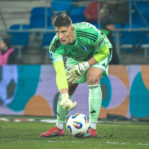 Bramkarz w zielonym stroju przygotowuje się do zagrania piłki w meczu piłki nożnej na stadionie.