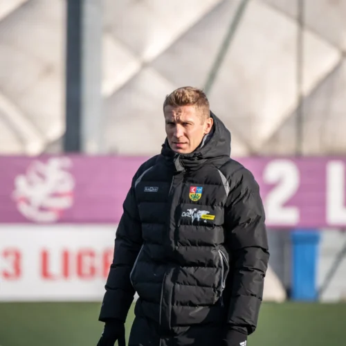 Na zdjęciu widać mężczyznę w ciemnej kurtce, stojącego na boisku piłkarskim, w tle widoczne są banery z napisami "2 LIGA" i "3 LIGA".