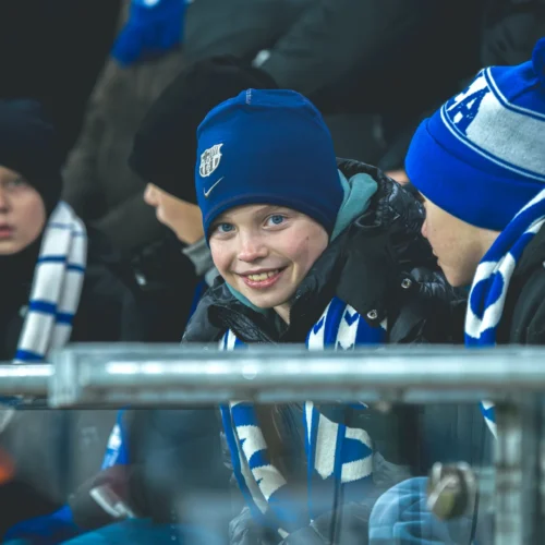 Na zdjęciu widać grupę dzieci siedzących na stadionie, z uśmiechniętym chłopcem w niebieskiej czapce i szaliku, cieszącym się atmosferą meczu.