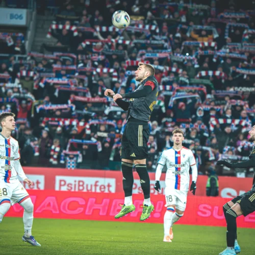 Na zdjęciu widać zawodników podczas meczu piłkarskiego, z jednym graczem skaczącym do piłki, a w tle kibice z transparentami.