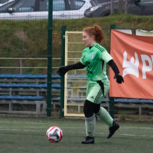 Kobieta w zielonym stroju bramkarskim gra w piłkę nożną na boisku z reklamą w tle.