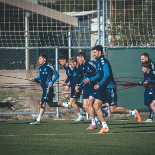 Na zdjęciu widać grupę piłkarzy trenujących na boisku, przywiązanych do siebie za pomocą kolorowych pasków, w słoneczny dzień.