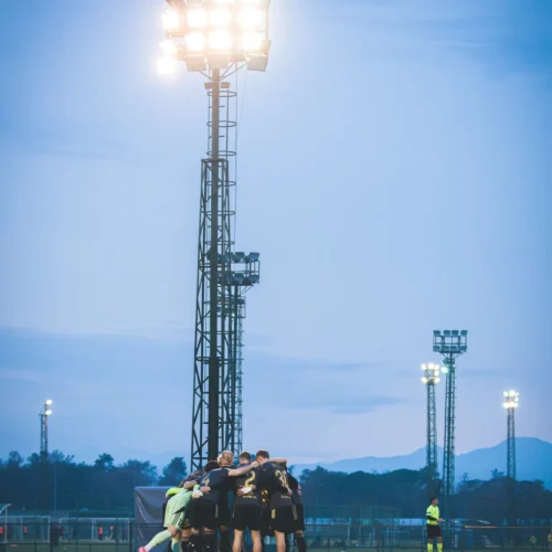Zespół piłkarski zbiera się w kręgu na boisku, oświetlonym przez potężne reflektory, w otoczeniu zmierzchu.