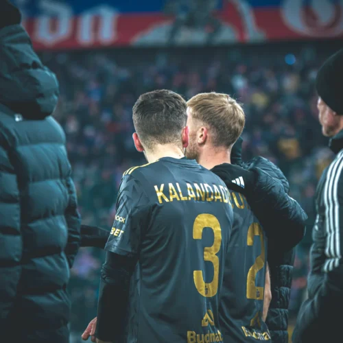 Zdjęcie przedstawia dwóch piłkarzy w czarnych koszulkach z numerami 3 i 2, stojących przy linii boiska podczas meczu, w towarzystwie innych osób w zimowych kurtkach.