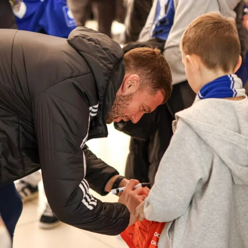 Na zdjęciu widoczny jest mężczyzna w czarnej kurtce, który podpisuje coś na ręce chłopca w szarej bluzie, otoczeni przez grupę ludzi.