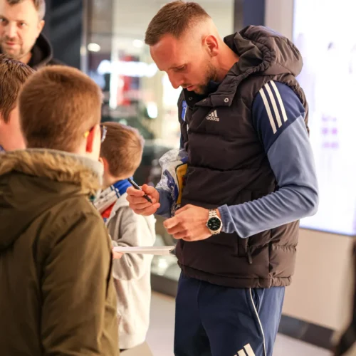 Na zdjęciu widoczny jest mężczyzna w sportowej odzieży, podpisujący autografy dla grupy dzieci w centrum handlowym.