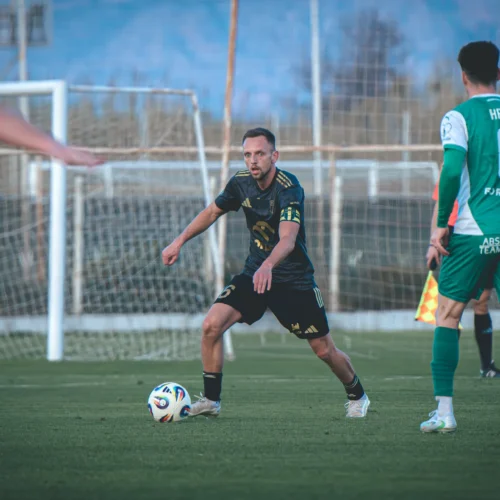 Na zdjęciu widoczny jest piłkarz w czarnym stroju, który przygotowuje się do zagraniu piłki na boisku, podczas gdy dwóch innych zawodników w zielonych strojach obserwuje sytuację.