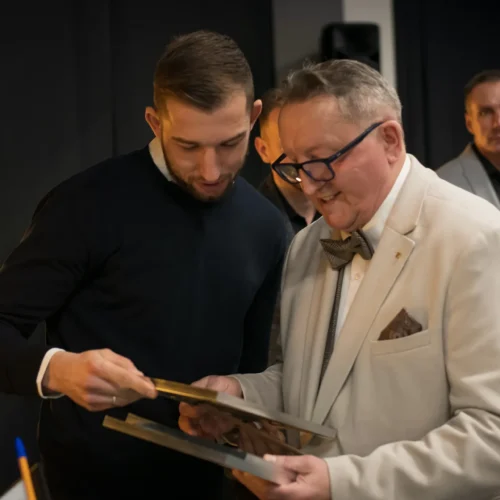 Na zdjęciu dwie osoby, jedna w jasnym garniturze, a druga w ciemnym swetrze, z fascynacją przeglądają zdjęcia lub dokumenty.