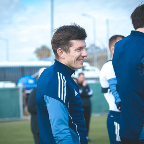 Na zdjęciu widać uśmiechniętego mężczyznę w sportowej odzieży, będącego w trakcie rozmowy z innymi osobami na boisku treningowym.