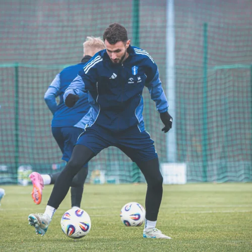 Na zdjęciu widać piłkarzy trenujących na boisku, z fokusem na zawodnika wykonującego zwód obok dwóch piłek.