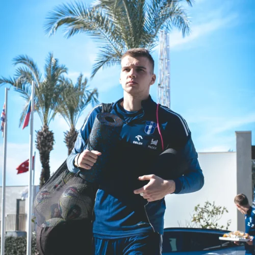 Mężczyzna w sportowym stroju niesie matę do ćwiczeń, przechodzi obok palm i flag w słoneczny dzień.