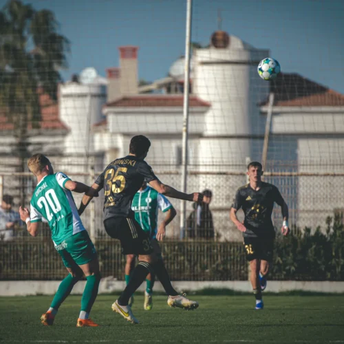 Na zdjęciu widać moment meczu piłkarskiego, w którym zawodnik w czarnej koszulce podbija piłkę, a jego przeciwnik w zielonym odwraca się do tyłu.
