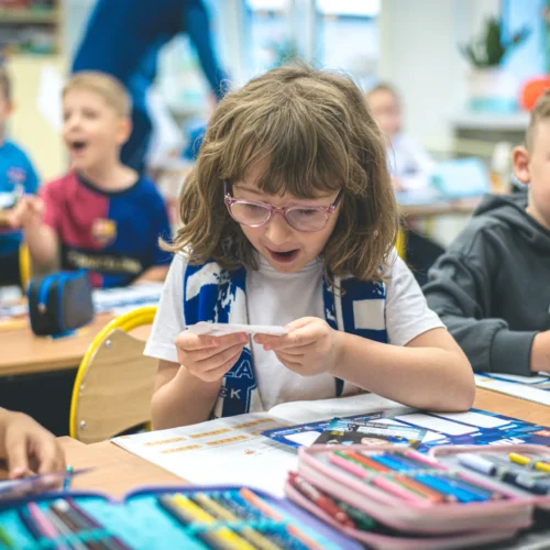Na zdjęciu widać uśmiechniętą dziewczynkę z okularami, która z zaciekawieniem patrzy na kartkę, podczas gdy w tle bawią się inne dzieci w klasie.