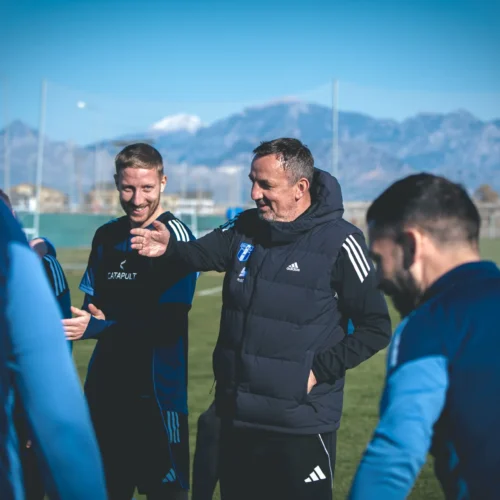 Zdjęcie przedstawia grupę mężczyzn w strojach sportowych, rozmawiających i śmiejących się na boisku treningowym w słoneczny dzień.