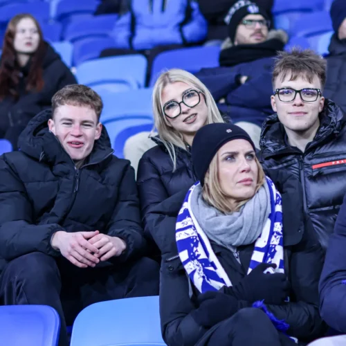 Na zdjęciu widoczna jest grupa ludzi, którzy siedzą na trybunach, ubrani w ciepłe kurtki, z wyraźnymi emocjami podczas wydarzenia sportowego.