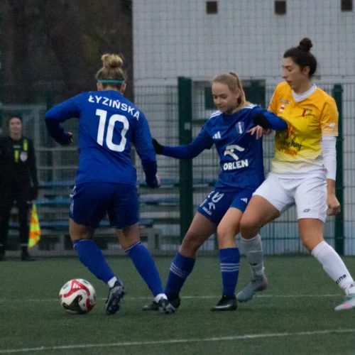 Na zdjęciu widać zawodniczki grające w piłkę nożną na boisku, z jedną z nich (Łyzińska, nr 10) próbującą przejąć piłkę od przeciwniczki.