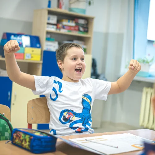 Chłopiec w białej koszulce z niebieskim wzorem radośnie podnosi ręce w geście zwycięstwa, siedząc przy biurku w klasie.