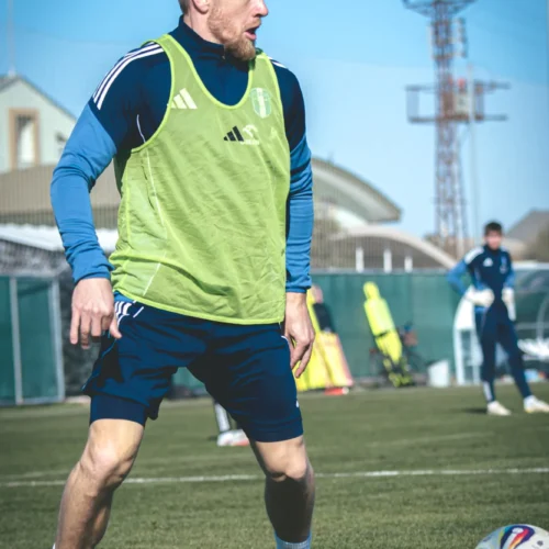 Piłkarz w zielonej kamizelce treningowej wykonuje ćwiczenia na boisku piłkarskim w słoneczny dzień.