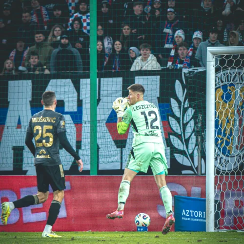 Na zdjęciu widać bramkarza w zielonym stroju, który uchwycił piłkę, podczas gdy zawodnik w czarnym uniformie, numer 25, porusza się w jego kierunku, a w tle znajdują się zaaferowani kibice.