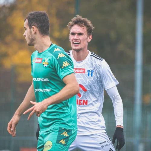 Na zdjęciu widać dwóch piłkarzy w trakcie meczu, przy czym jeden z nich wyraża silne emocje, a drugi zwraca uwagę na sytuację.