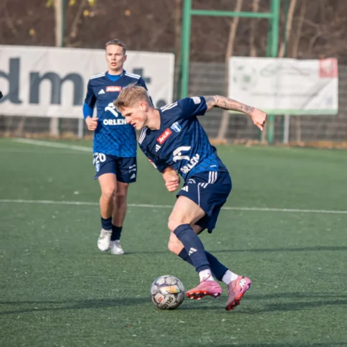 Na boisku piłkarskim dwóch zawodników w niebieskich strojach rywalizuje z zawodnikiem w białym dresie, a piłka znajduje się w trakcie akcji.