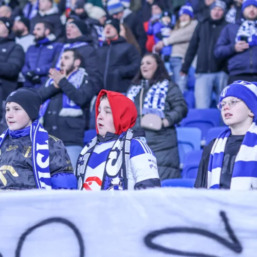 Na zdjęciu widoczni są trzej młodzi kibice w szalikach, śpiewający na trybunach podczas meczu sportowego.