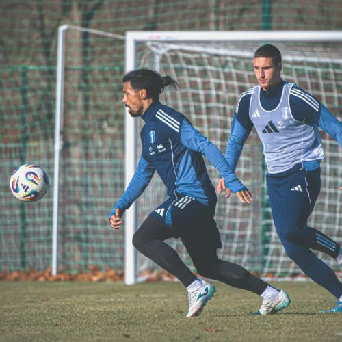 Dwa piłkarze trenują na boisku, z piłką w powietrzu w trakcie sesji treningowej.