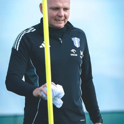 Mężczyzna w czarnej odzieży sportowej trzyma żółtą tyczkę i materiał w ręku, spacerując po boisku treningowym.