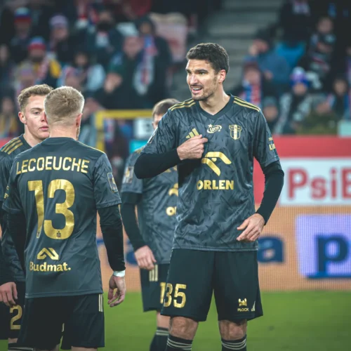 Na zdjęciu widać grupę zawodników w piłkarskich strojach, z numerami na plecach, rozmawiających ze sobą podczas meczu.