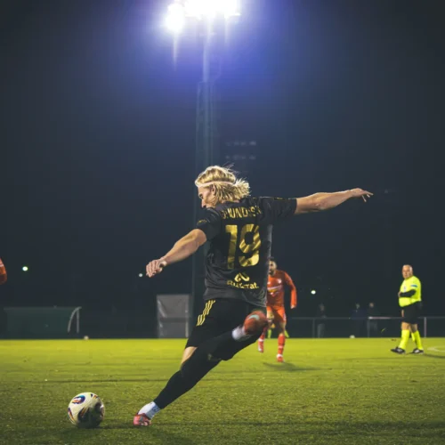 Na zdjęciu widać piłkarza w czarnej koszulce z numerem 19, który wykonuje strzał na bramkę w czasie nocnego meczu piłkarskiego, a w tle znajdują się zawodnicy w pomarańczowych strojach.