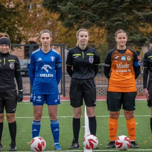 Na zdjęciu widocznych jest pięć osób w strojach piłkarskich na boisku, w tym sędziowie oraz zawodniczki, stojące obok siebie z piłkami u stóp.