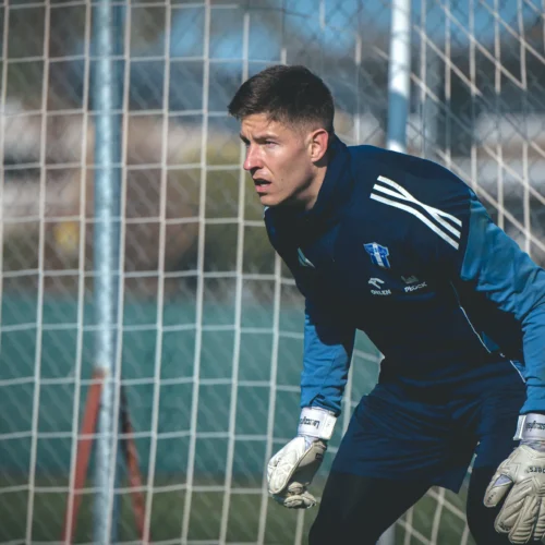 Młody bramkarz w niebieskim stroju sportowym stoi w pozycji gotowej do interwencji w piłkarskiej bramce.