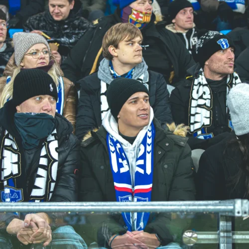 Grupa kibiców w odzieży zimowej siedzi na trybunach, kibicując swojej drużynie sportowej.
