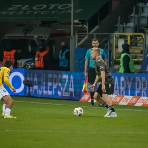 Na zdjęciu przedstawiono zawodników podczas meczu piłkarskiego na boisku, gdzie jeden z nich przygotowuje się do zagrania piłki.