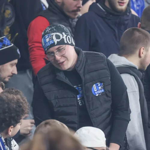 Na zdjęciu widoczna jest grupa kibiców, z wyraźnym fokusem na młodego mężczyznę w czarnym kapeluszu, który rozmawia i uśmiecha się do innych fanów.