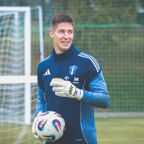 Na zdjęciu widoczny jest bramkarz w stroju sportowym, trzymający piłkę, stojący na boisku w pobliżu bramki.