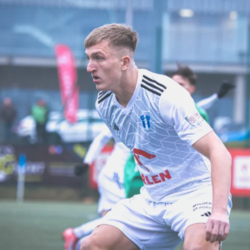 Młody piłkarz w białej koszulce biega po boisku w trakcie meczu.