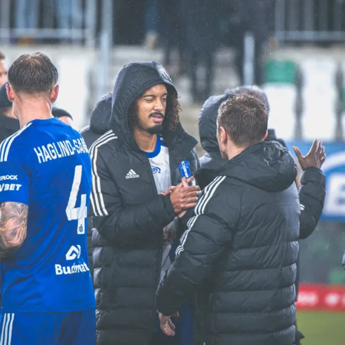 Na zdjęciu widoczna jest scena z meczu piłkarskiego, gdzie zawodnicy w niebieskich strojach i sztab szkoleniowy wymieniają uściski rąk w deszczowej atmosferze.