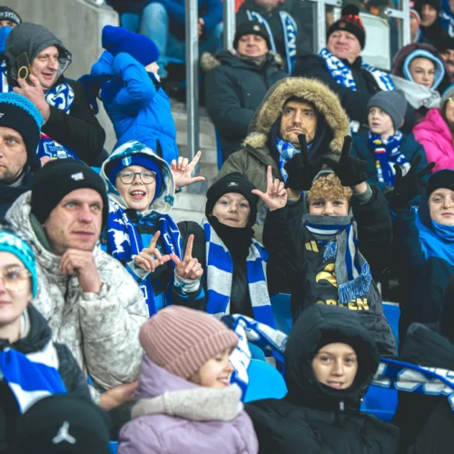 Widok tłumu kibiców na stadionie, po części w strojach zimowych, wśród których znajdują się radosne dzieci z szalikami w barwach drużyny.