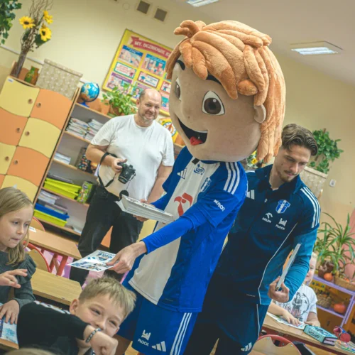 W klasie uczniowie uśmiechają się, podczas gdy jedna osoba w kostiumie maskotki z dużą, blond fryzurą rozdaje im materiały do nauki.