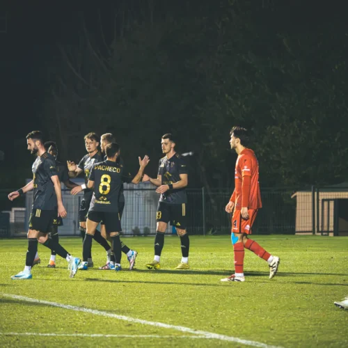 Na zdjęciu widać grupę piłkarzy cieszących się po zdobyciu bramki, z jednym zawodnikiem w czerwonym stroju jako bramkarzem w tle.