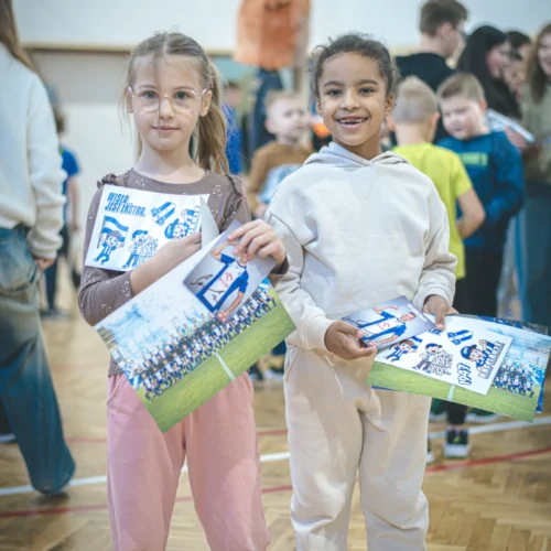 Dwie dziewczynki uśmiechają się, trzymając w rękach kolorowe ulotki w zatłoczonej sali.