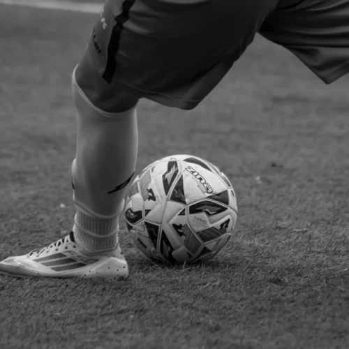 Na zdjęciu w czarno-białej tonacji widać nogę piłkarza stojącego obok piłki na trawie.