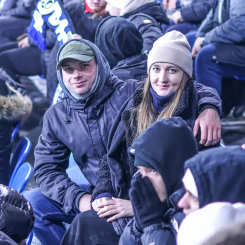 Na zdjęciu widać młodą parę siedzącą na trybunach stadionu, otoczoną innymi kibicami, w zimowych ubraniach.